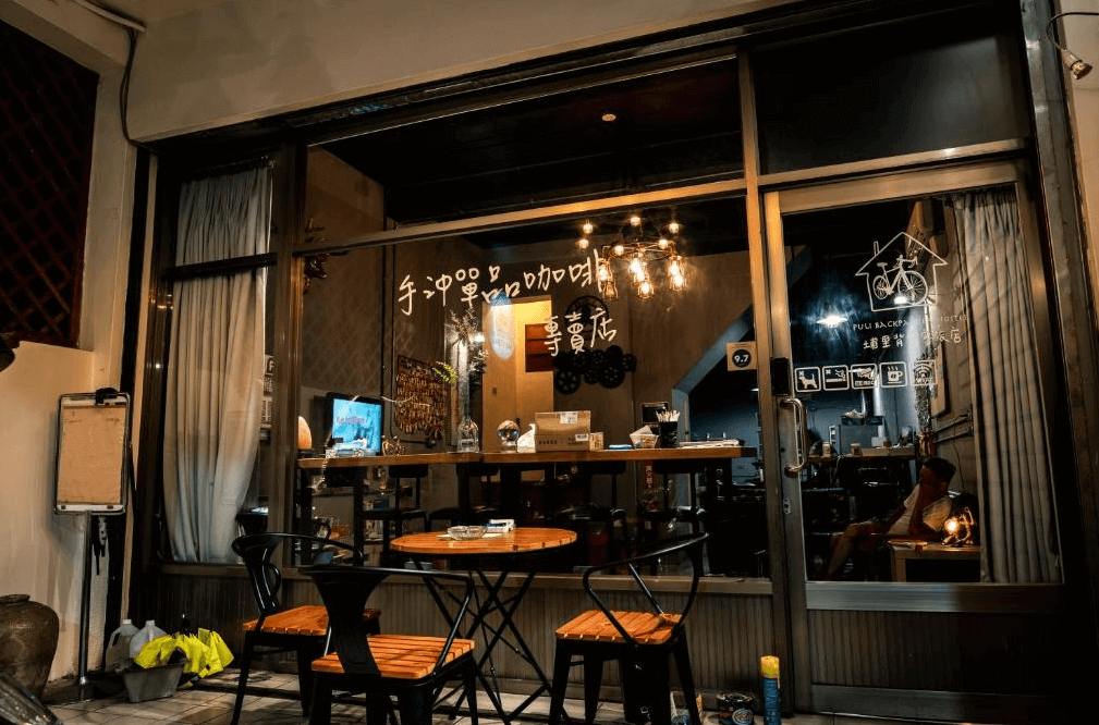 【埔里住宿】麋路背包客旅店外觀的夜景，玻璃窗內透出溫暖燈光，店內設有手沖咖啡專區，搭配簡約木桌與椅子，營造舒適的社交氛圍。