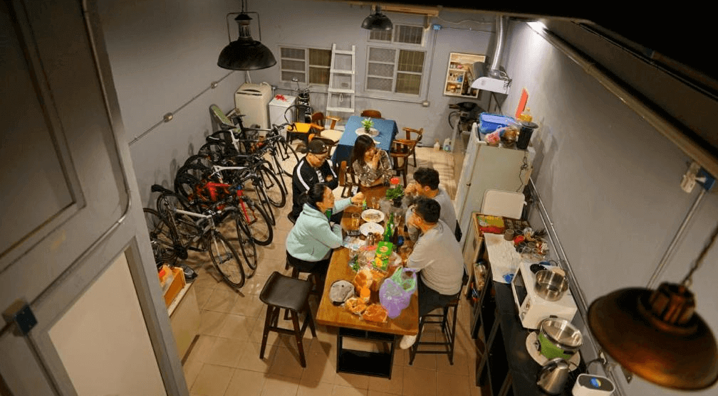 【埔里住宿】麋路背包客的公共空間，中心擺放長木桌，數人圍桌用餐或交流。背景可見存放的腳踏車和廚房設備，提供實用性與居家感。