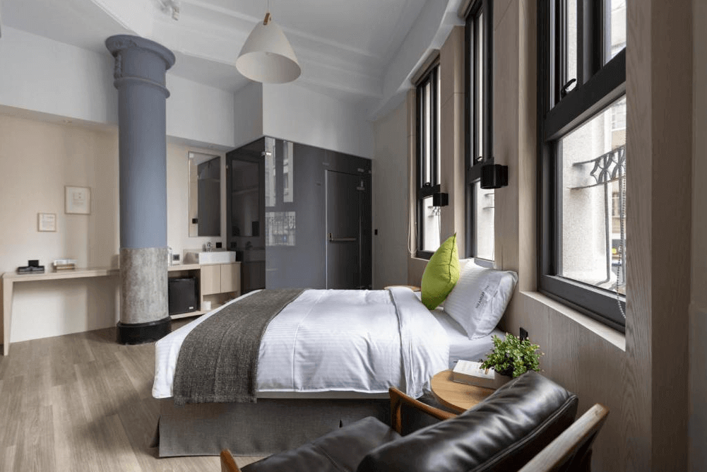 【台北車站住宿】"台北車站青年旅館"MEANDER 1948的復古工業風雙人房，設有一張舒適大床，搭配高挑天花板與復古柱體設計。房內擁有大面積窗戶，採光充足，並設有木質書桌、皮革扶手椅與簡約的衛浴空間，展現現代與復古融合的美感。