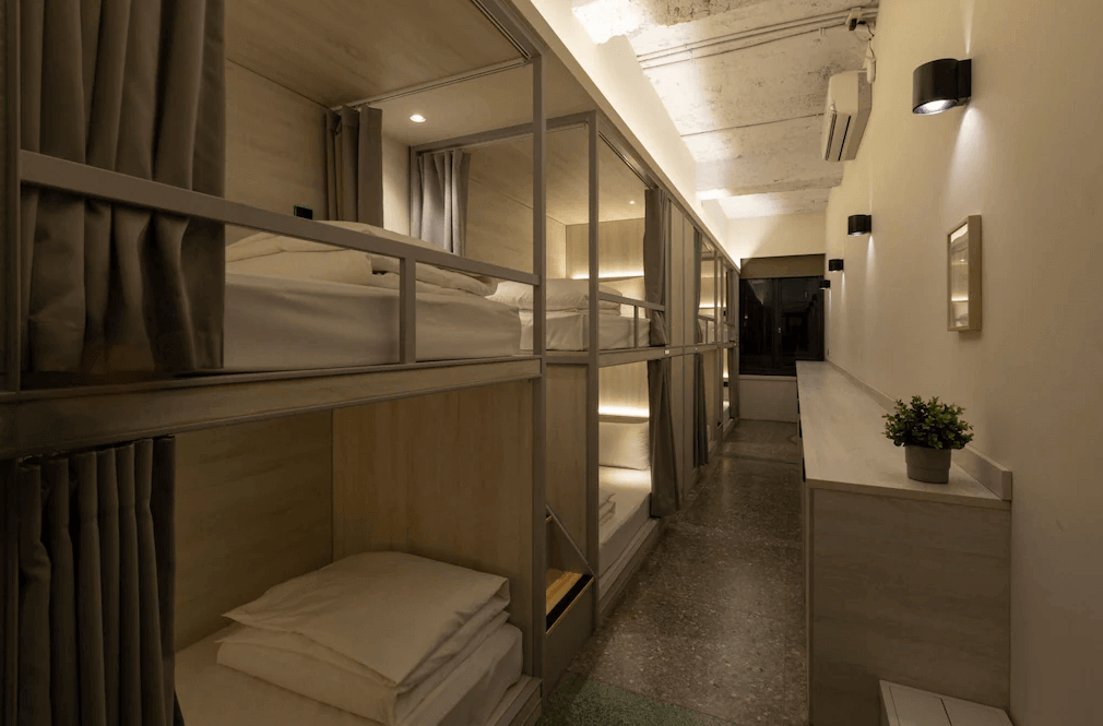 【台北車站住宿】"台北車站青年旅館"MEANDER 1948的宿舍房型，擁有多個上下舖床位，每個床位皆附設隱私遮簾與個人閱讀燈。室內燈光柔和，牆面搭配簡約植栽擺飾，營造舒適共享空間。