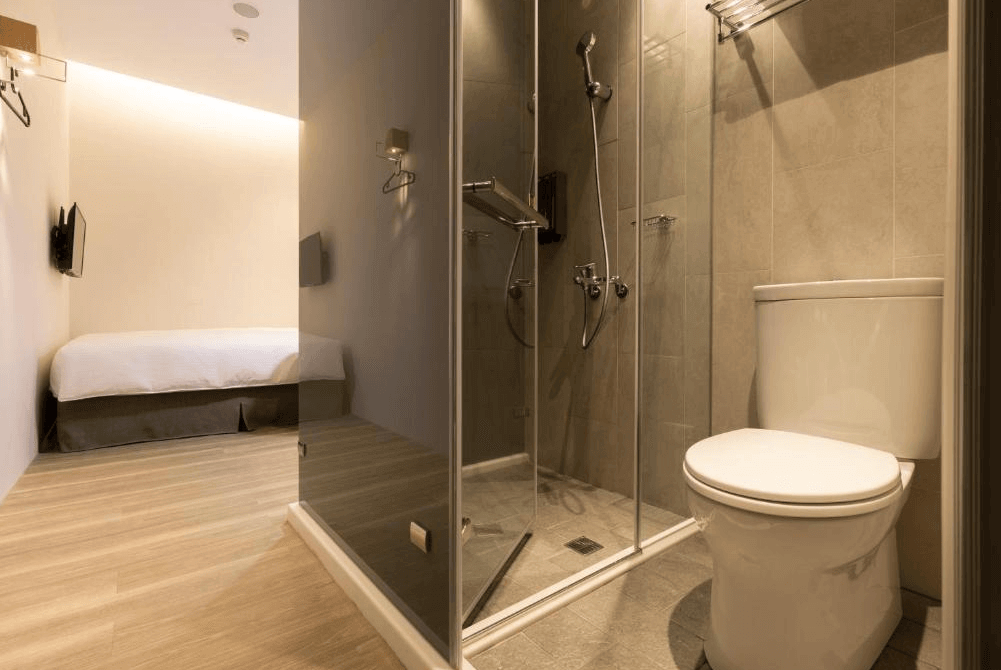 【台北車站住宿】"台北車站青年旅館"MEANDER 1948的私人衛浴空間，採用玻璃淋浴間設計，搭配灰色瓷磚牆面與白色洗手台。空間規劃簡約且具功能性，提供乾淨舒適的住宿體驗。