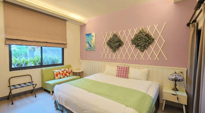 倍詩泰民宿溫馨浪漫的雙人房，牆面刷上淡紫色並搭配木質裝飾，床頭點綴著藤編花架與綠色植栽，營造清新自然的氛圍。大床搭配柔軟的綠色床巾與格紋抱枕，窗邊擺放著小型沙發，增添舒適感。