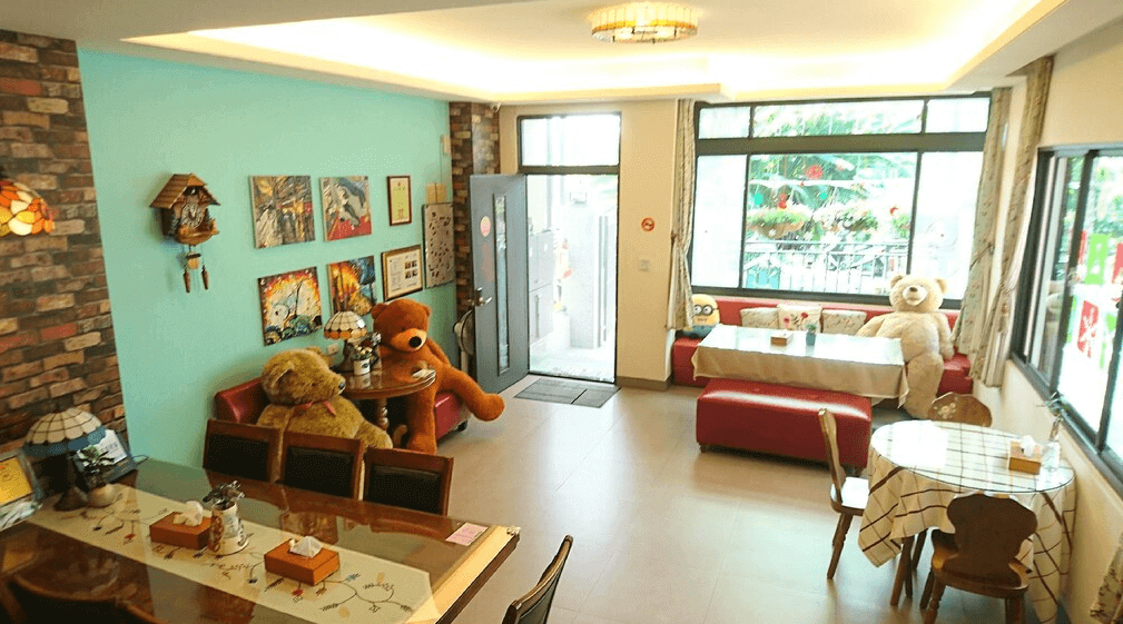 【集集火車站住宿】倍詩泰民宿的公共空間，採用亮眼的湖水綠牆面搭配磚牆裝飾，空間內設有多張座椅與長桌，擺放著可愛的大型泰迪熊，牆上則點綴著溫馨掛畫與擺飾，營造出溫暖且富有童趣的用餐與交流空間。