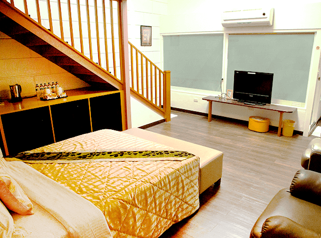 【四重溪溫泉】優生美地休閒農場樓中樓設計的家庭房，房內設有一張大床，搭配金黃色的寢具與米色床頭靠墊，營造溫暖舒適的氛圍。房間內有木質樓梯通往閣樓，提供更多睡眠空間。設備包括電視、空調、皮沙發與木質收納櫃，空間寬敞適合家庭入住。