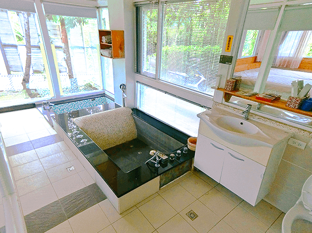 【四重溪溫泉】優生美地休閒農場的泡湯房，內部設有黑色石砌浴池，旁邊擺放石製靠枕，提供舒適泡湯體驗。浴池緊鄰大面積落地窗，可欣賞戶外綠意景色，增添放鬆感。房內另設有洗手台與白色收納櫃，維持整潔實用的空間。