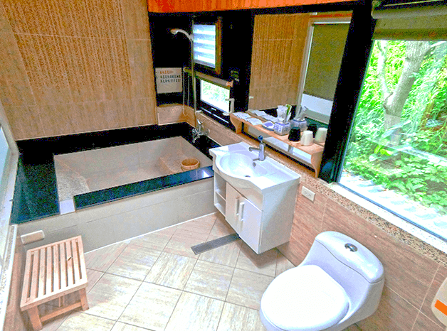 【四重溪溫泉】優生美地休閒農場木質與石材裝潢的溫泉湯屋，浴池以黑色石材打造，周圍設有木質長椅與窗邊綠意景觀，營造自然放鬆氛圍。洗手台搭配白色櫃體，窗邊設有開放式收納架，擺放盥洗用品，整體設計兼具舒適與實用。