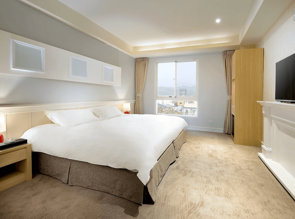 【集集火車站住宿】卡菲爾花園飯店典雅的雙人房，配有一張超大床，床鋪潔白柔軟，搭配溫暖的米色地毯與淡灰色牆面，營造舒適高雅的氛圍。房內設有大片窗戶，可欣賞遠方山景，自然光線充足，讓房間更加明亮開闊。