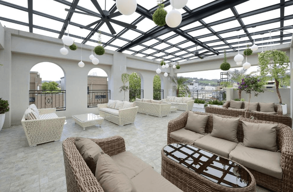 卡菲爾花園飯店歐式風格的露天休憩區，設有多組編織沙發與玻璃茶几，搭配綠意盎然的植栽與懸掛燈飾，打造典雅舒適的戶外空間。屋頂採透明玻璃設計，可享受陽光灑落的溫暖感，適合旅客放鬆與交流。