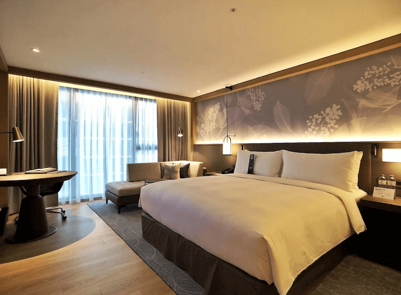 【台北車站住宿】台北中山九昱希爾頓逸林酒店豪華現代風格的酒店客房，設有一張大床，床頭牆面裝飾有優雅的花葉圖案。房內採用暖色調照明，配備落地窗、沙發椅、辦公桌和閱讀燈，營造舒適溫馨的氛圍。