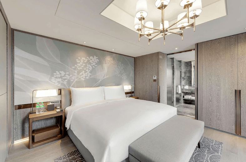 【台北車站住宿】台北中山九昱希爾頓逸林酒店明亮寬敞的酒店客房，擁有一張大床與灰藍色系花葉壁紙，搭配高級吊燈與木質家具，營造現代優雅的居住空間。房內擁有開放式設計，可直通豪華大理石浴室。