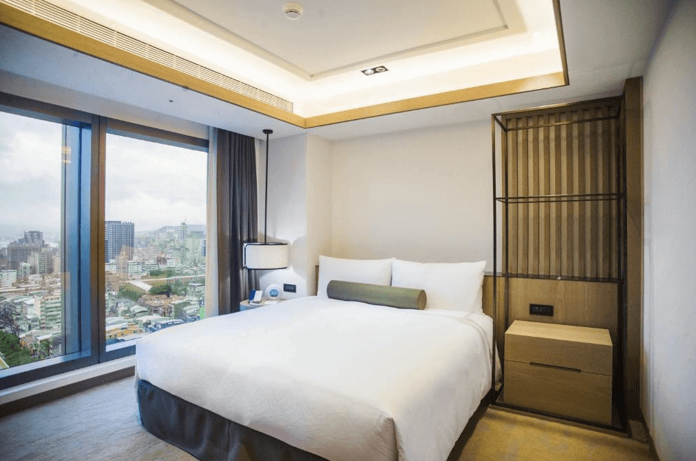 【台北親子飯店】台北凱達大飯店的高樓層雙人房，設計簡約大方，採用木質與暖色調搭配，營造溫馨舒適的氛圍。大面積落地窗提供開闊的城市景觀，床邊設有懸掛式燈具與床頭櫃，展現現代感與實用性兼具的住宿環境。