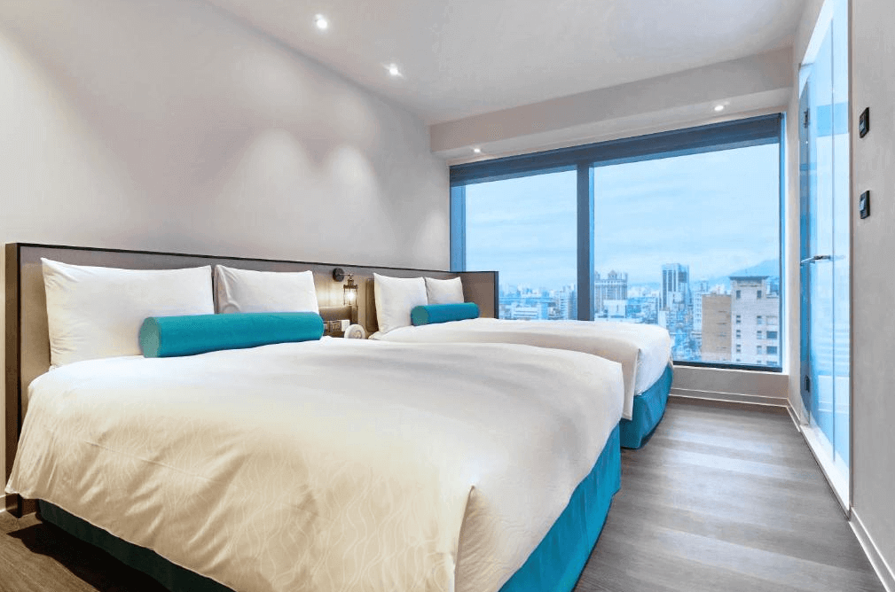 【台北親子飯店】台北凱達大飯店的四人房，設有兩張大床，適合家庭或團體入住。房內以藍白色系為主，並配有藍色長抱枕點綴。落地窗設計讓房間擁有充足的自然光，窗外可俯瞰台北市區美景，提供寬敞舒適的住宿體驗。