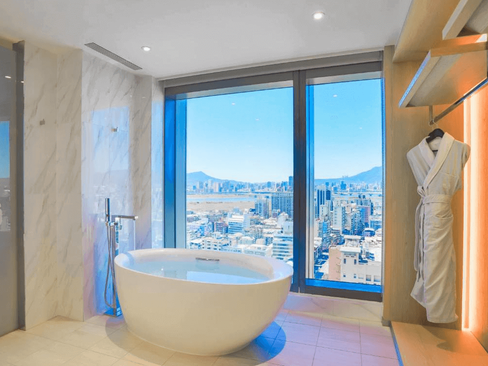 【台北親子飯店】台北凱達大飯店的景觀浴室，設有獨立白色浴缸，搭配大面積落地窗，可俯瞰台北市區與遠山景色。浴室內部採用大理石牆面與木質元素，提供高級飯店的典雅與舒適感，讓旅客在泡澡時享受放鬆時光。