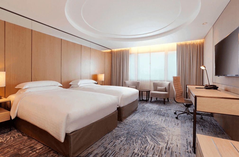 "台北親子飯店"【台北車站住宿】台北喜來登大飯店雙床客房，房內設有兩張舒適的單人床，搭配木質牆面與暖色調燈光，呈現簡約現代風格。窗邊設有兩張休閒椅與小桌，另配備辦公桌與人體工學椅，提供舒適的住宿體驗。
