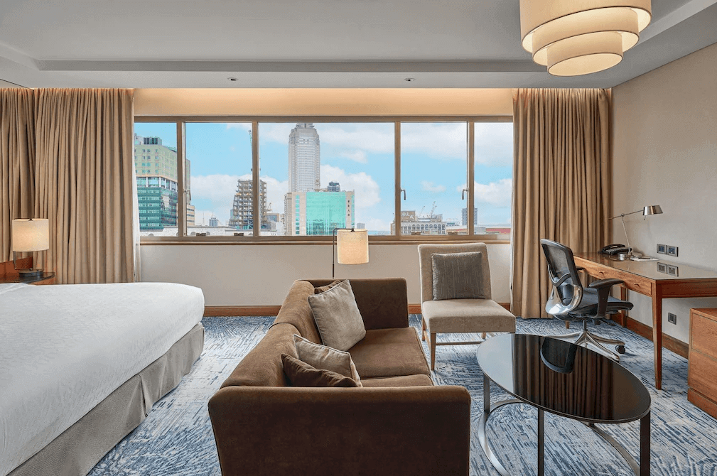 "台北親子飯店"【台北車站住宿】台北喜來登大飯店的豪華高樓層客房，設有一張大床與寬敞的窗景，俯瞰台北市區。房內配置沙發、玻璃茶几與辦公空間，融合現代設計與舒適機能，並以暖色調窗簾與燈光營造溫馨氛圍。