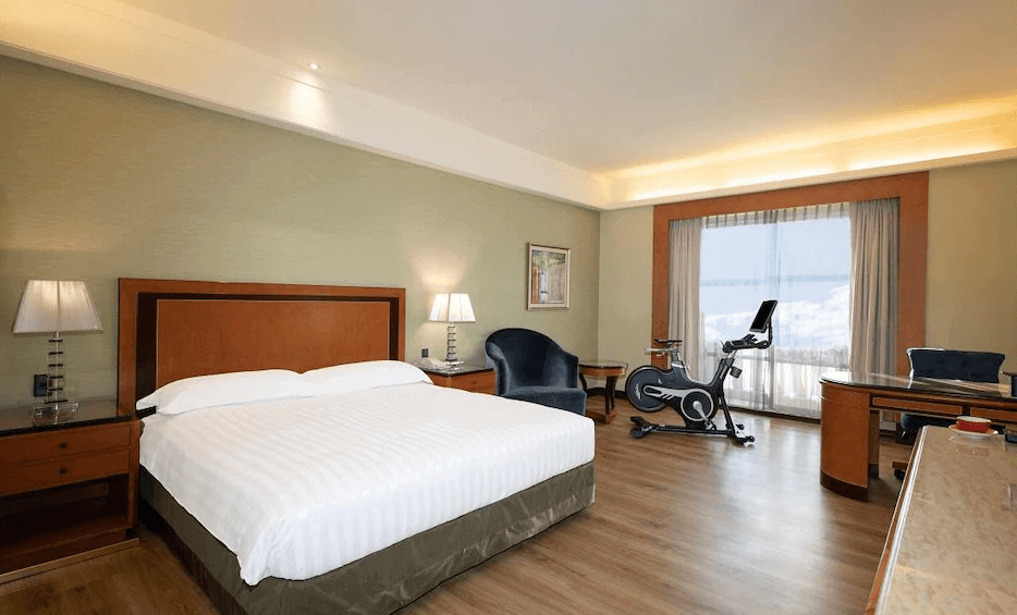 【松山機場住宿】「台北漢普頓酒店」客房內部，設有一張大床，房內配有木質家具、溫暖燈光與簡約裝潢。房間內還設有運動健身腳踏車、大型書桌與休閒椅，適合商務旅客與健身愛好者。