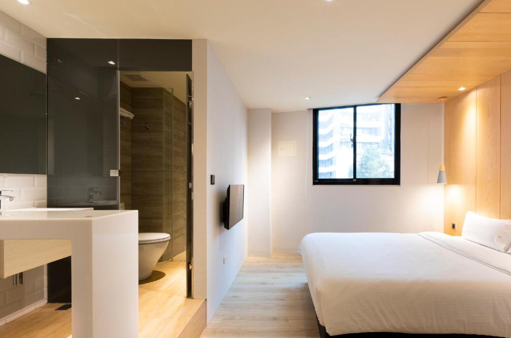 【西門町住宿】台北漫步旅店的客房，整體色調以白色和木質元素為主，搭配大面窗戶提供自然採光。開放式浴室設計，包含玻璃隔間的淋浴間和獨立洗手台，空間感寬敞舒適。