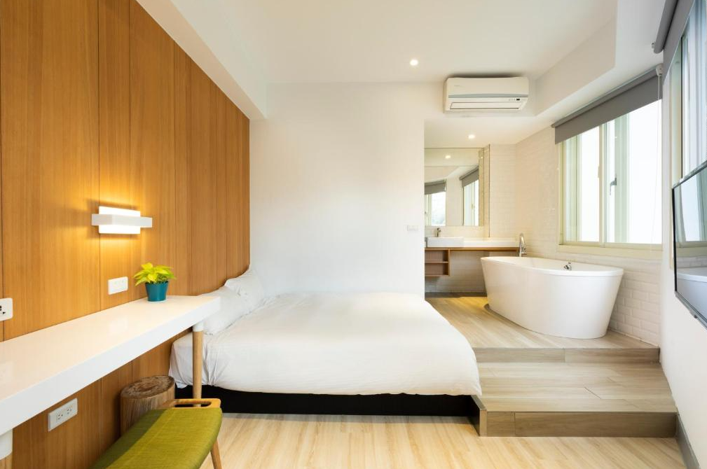 【西門町住宿】台北漫步旅店的雙人房，木質牆面與白色家具營造溫暖氛圍，房內設有一張雙人床、壁燈、簡約桌椅，並配有開放式浴室區，帶有獨立浴缸，適合享受放鬆的住宿體驗。