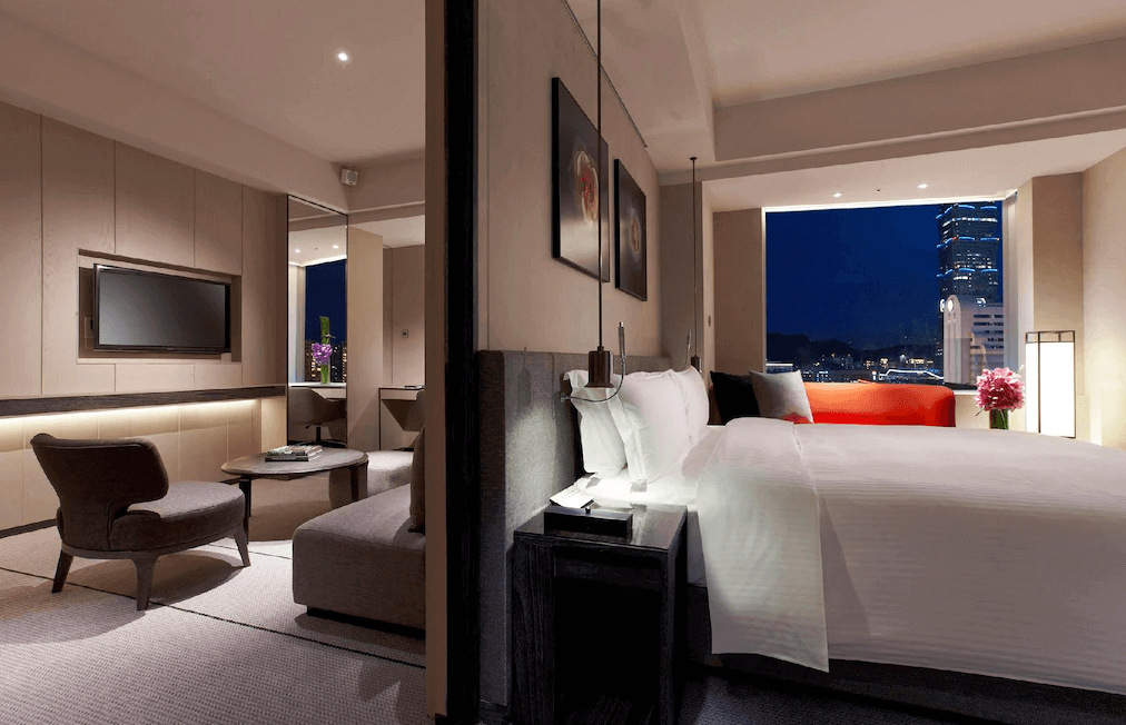 【台北親子飯店】台北艾麗酒店的豪華套房，臥室內擁有一張大床，搭配床邊吊燈與夜景窗景。客廳區設有沙發與茶几，提供寬敞的休憩空間。透過窗戶可見台北101大樓的壯麗夜景，增添住宿的奢華感。