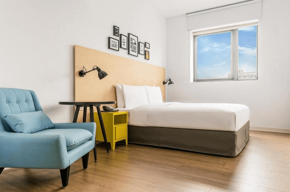 台北西門町意舍酒店的雙人房，搭配現代設計風格。房內擺放一張大床，木質床頭牆面上裝飾著多幅黑白畫作，並設有壁掛式閱讀燈。角落配置藍色單人沙發與抱枕，黃黑相間的床頭櫃與圓形邊桌增添活潑感，大片窗戶帶來充足自然光。