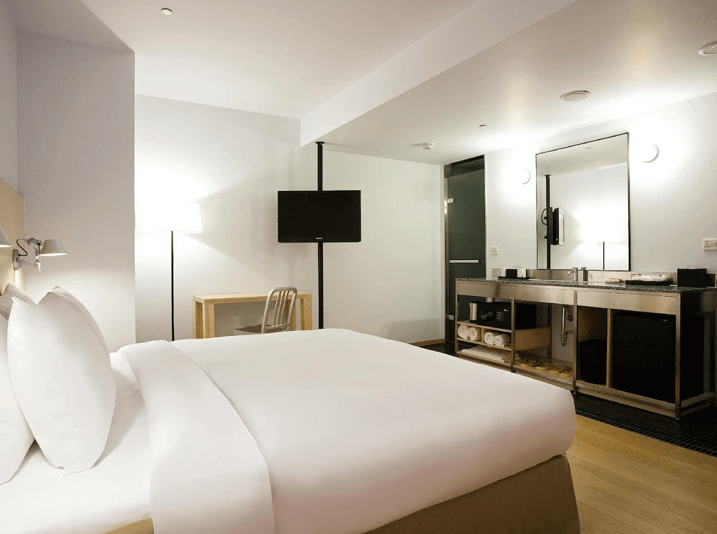 台北西門町意舍酒店的雙人房，採用簡約的白色床鋪，床邊裝有壁燈，提供溫暖的光線。房間內設有開放式洗手台，鋼架設計搭配黑色檯面，下方擺放毛巾與基本備品。懸掛式電視與金屬材質的書桌椅，讓空間更具現代感。