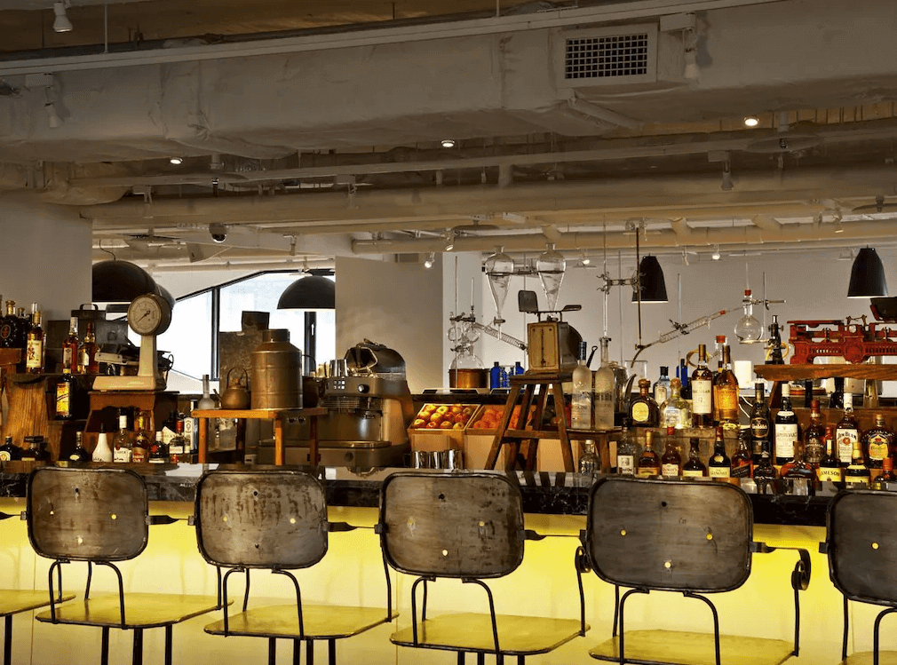 台北西門町意舍酒店的復古與工業風格酒吧，櫃檯前擺放金屬復古高腳椅，背景為開放式酒架，陳列各式烈酒與調酒器具。天花板採用裸露管線設計，搭配懸掛式燈具，營造獨特的都市氛圍。