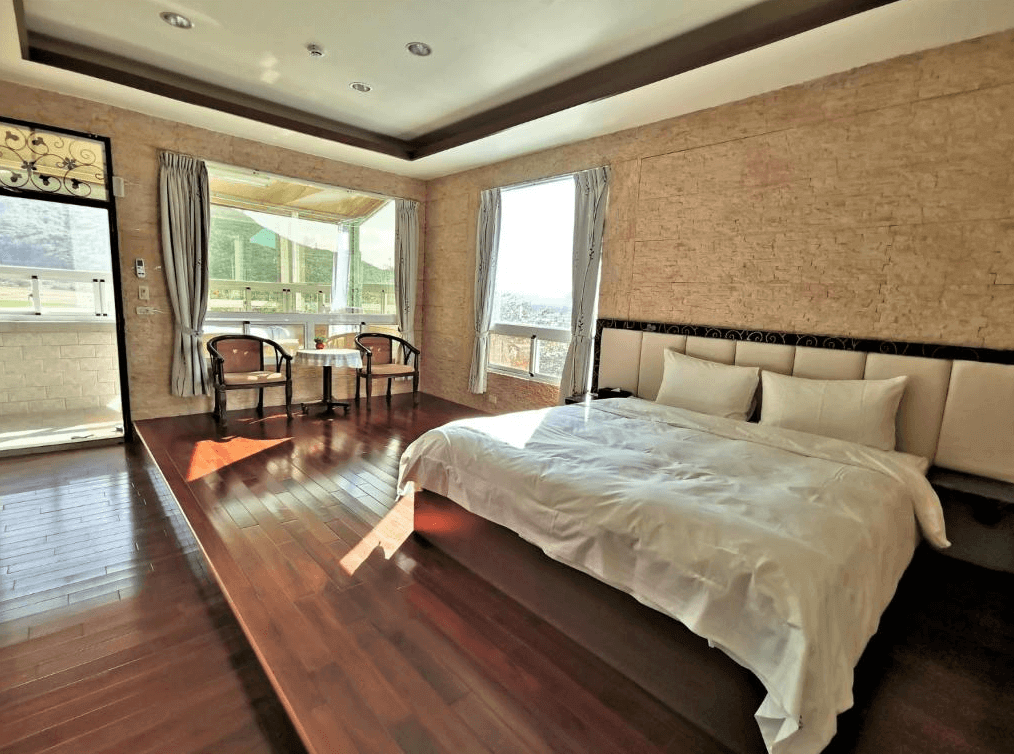 【四重溪溫泉】四重溪可以居民宿的明亮客房，設有一張大床，搭配米色軟墊床頭與淺色石紋牆面，呈現自然簡約風格。深色木質地板與挑高設計讓空間感更顯寬敞。窗邊擺有一組木製桌椅，可在此享受窗外青山美景與溫暖日光，營造悠閒舒適的住宿體驗。