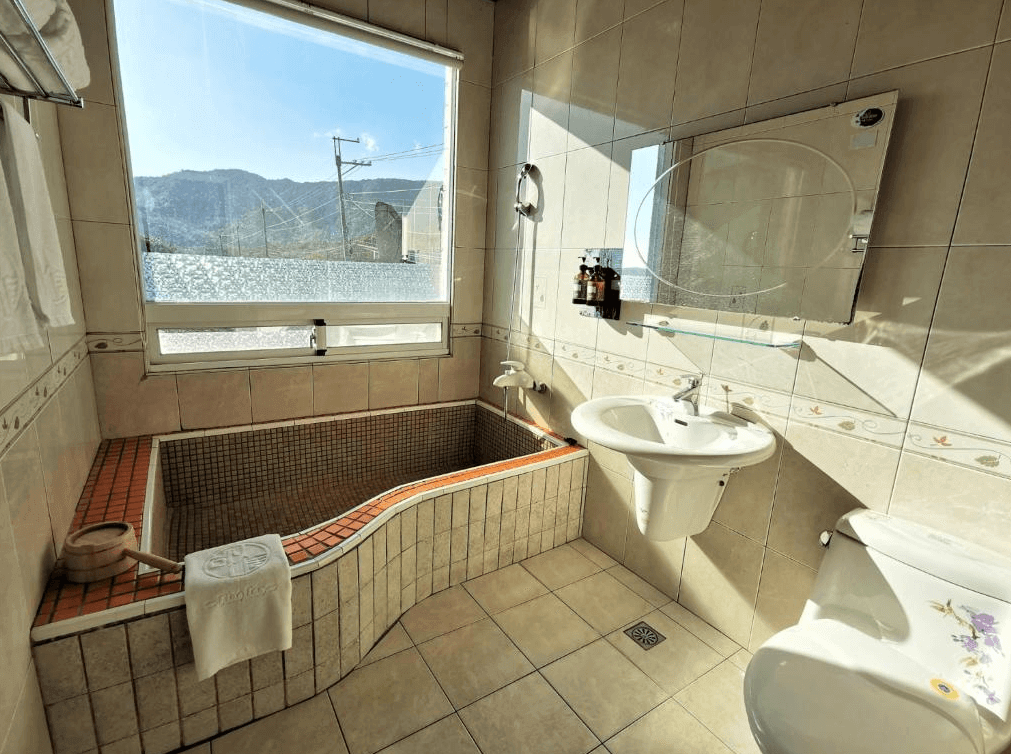 【四重溪溫泉】四重溪可以居民宿傳統日式設計的浴室，內設獨立泡湯池，池邊使用紅色與米色馬賽克磁磚鋪設，帶來溫暖的視覺效果。窗戶提供自然光線，可在泡湯時欣賞戶外山景，營造放鬆愜意的氛圍。浴室內配備白色洗手台與鏡子，整體設計乾淨整潔。