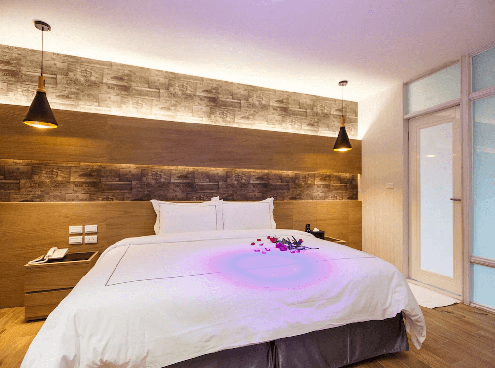 【金山溫泉】大埔硫磺溫泉商旅的客房，配有大型雙人床，床上撒有玫瑰花瓣。房間採用木質與磚紋牆面設計，並搭配暖色吊燈與間接照明，營造出舒適的氛圍。旁邊有木質床頭櫃與電話，房間整潔且佈置雅緻。