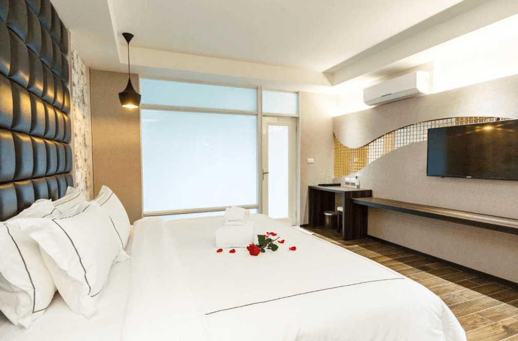 【金山溫泉】大埔硫磺溫泉商旅的高級客房，擁有大床、皮革質感的床頭板與白色枕被，床上鋪有毛巾和玫瑰花瓣。房內設有懸掛式平面電視、長桌與洗手台區，並搭配大面積的玻璃窗，引入充足的自然光，讓空間更加明亮開闊。