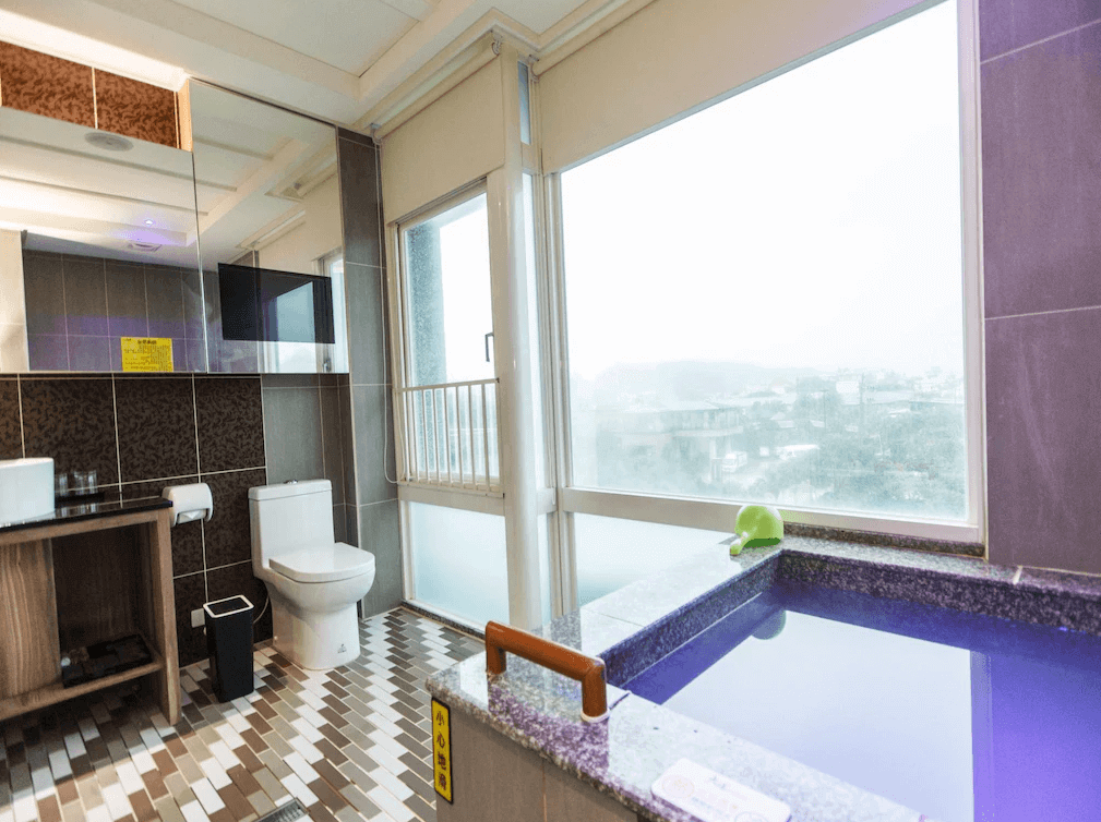 【金山溫泉】大埔硫磺溫泉商旅帶有景觀的大理石溫泉浴池，位於衛浴空間內，搭配落地窗可欣賞戶外山景或市景。浴室設有馬桶、洗手台與懸掛式鏡面電視，地面採用拼接瓷磚，整體風格現代且典雅。