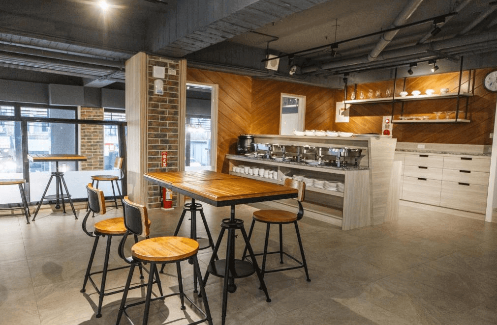 【金山溫泉】大埔硫磺溫泉商旅的自助餐廳，擁有工業風設計，搭配木質高腳桌椅與磚牆裝飾。餐檯上擺放著多個餐盤與自助餐設備，空間明亮且整潔，提供舒適的用餐環境。