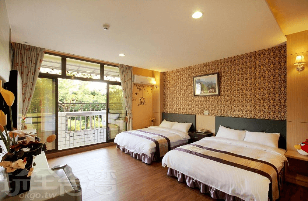 【集集火車站住宿】富麗渡假民宿典雅寬敞的四人房，設有兩張舒適的雙人床，床頭牆面採用華麗的花紋壁紙裝飾，增添質感。大片落地窗提供明亮自然光，可通往陽台，欣賞戶外綠意景觀，搭配暖色調燈光營造溫馨氛圍。