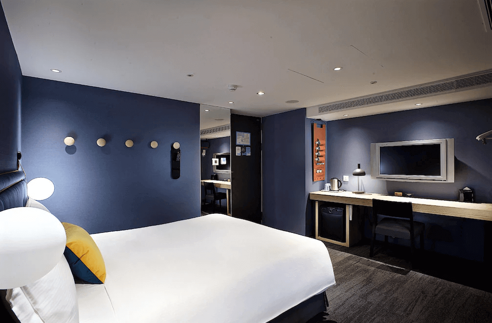 【西門町住宿】捷絲旅台北西門館的客房，主色調為深藍色，配有一張雙人床，牆上有裝飾掛鉤與壁燈，書桌區域設有電視、迷你冰箱和熱水壺，空間設計簡約且實用。