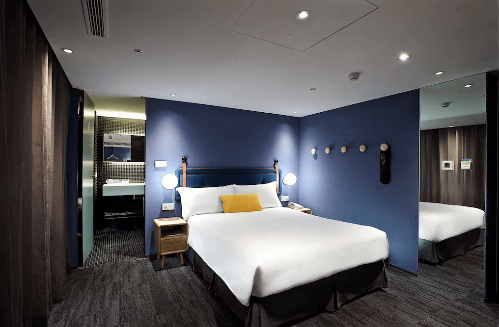 【西門町住宿】捷絲旅台北西門館的客房，牆面為深藍色，床頭燈具別具設計感，搭配黃藍色抱枕，浴室區域採開放式設計，洗手台與衣架區以玻璃隔開，營造出時尚氛圍。