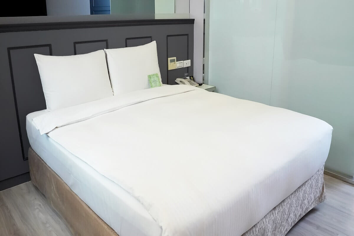 【西門町住宿】旅居文旅西門驛站-西寧館的客房，擺放著一張整潔的雙人床，床鋪鋪著純白色床單與兩個白色枕頭。床頭背板為深灰色設計，牆邊設有插座和電話，床邊有玻璃隔間，隱約可見浴室空間。