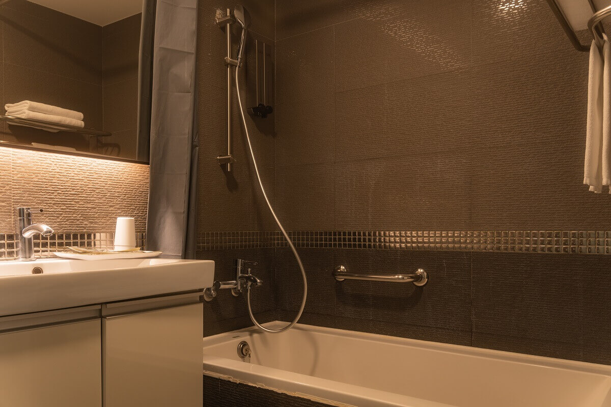 【西門町住宿】旅居文旅西門驛站-西寧館的浴室，牆面採用黑色與棕色馬賽克瓷磚，內有一個浴缸，配備可調式蓮蓬頭與壁掛式電熱水器。洗手台旁擺放著盥洗用品，鏡面上方設有間接照明，整體氛圍溫暖且具有質感。