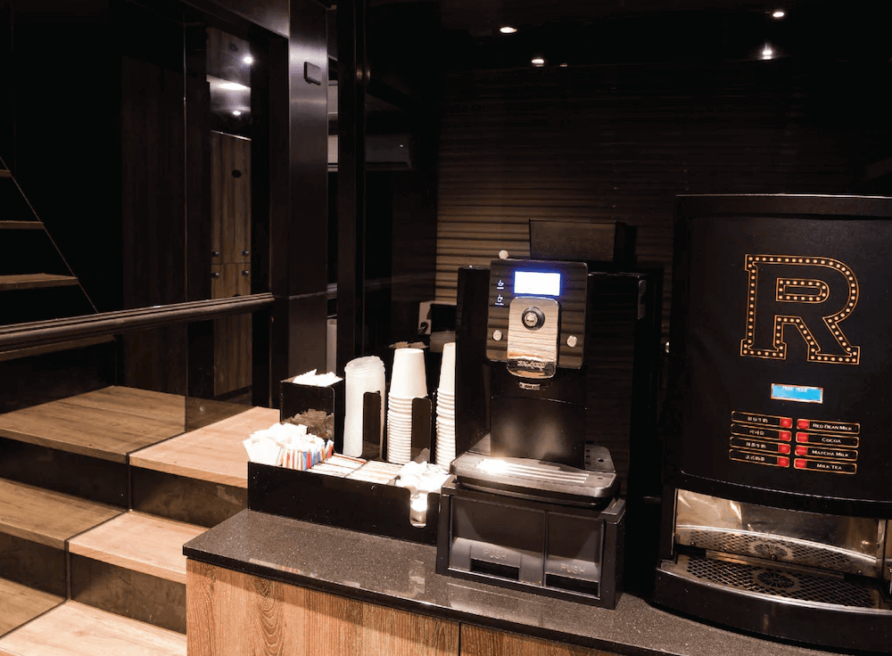 【台北車站住宿】（旅樂序精品旅館 站前五館）：自助咖啡吧，提供免費飲品與咖啡機，擺放多種飲品選項與即飲杯，供住客自行取用。整體空間採用黑色與木質調設計，展現時尚氛圍。