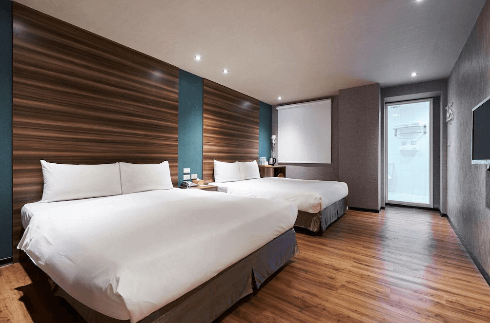 【西門町住宿】明日大飯店寬敞的客房，內部設有兩張大床，搭配木質地板與深色木紋牆面，燈光溫暖且柔和。房內設有獨立浴室，浴室門為透明玻璃設計，整體風格現代且舒適