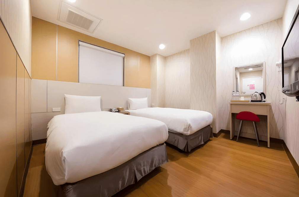 【西門町住宿】明日大飯店的雙床房，兩張單人床擺放整齊，房間色調為溫暖的米色與淺木色，搭配簡約的裝潢與木質地板。房內設有梳妝台、小型書桌與紅色椅凳，燈光明亮且溫馨。
