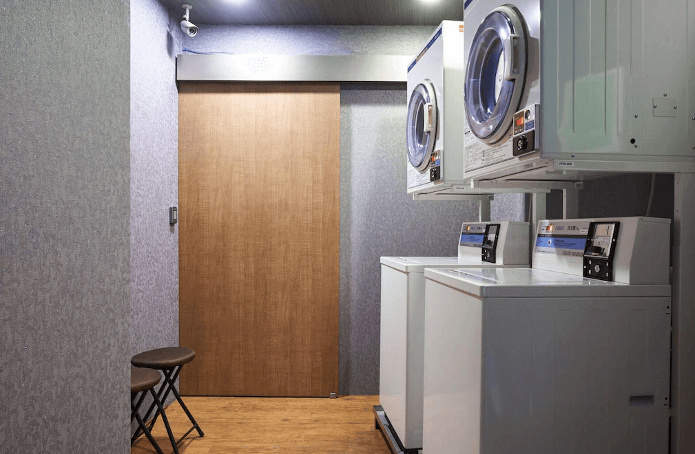 【西門町住宿】明日大飯店的自助洗衣空間，內有多台洗衣機與烘衣機，設備現代且整齊擺放。洗衣區內部裝潢簡約，牆面為深灰色調，並設有小型折疊椅供使用者休息。