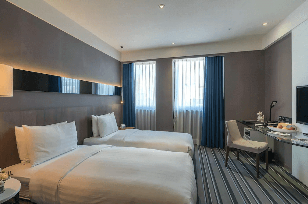 【西門町住宿】永安棧的雙床房，兩張單人床鋪著純白色床單，床頭牆面為深灰色，搭配間接照明設計。房間內有大片窗戶，藍色窗簾增添沉穩氛圍，並配置玻璃書桌與椅子，適合商務或休閒旅客入住。