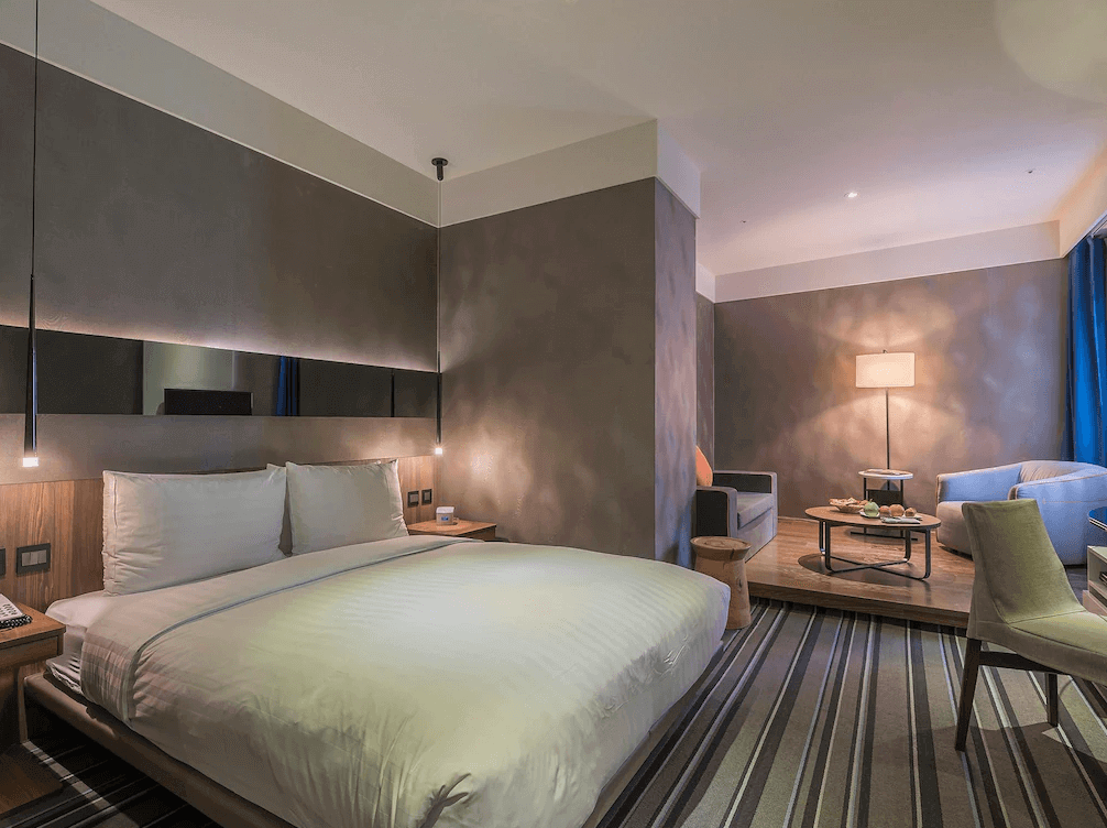 【西門町住宿】永安棧的雙人房，擺放一張大床，床頭牆面採用灰色塗層設計，搭配懸吊式吊燈增添現代感。房內設有小型起居區，擺放沙發與茶几，營造舒適的休憩空間，並以暖色系燈光提升整體氛圍。