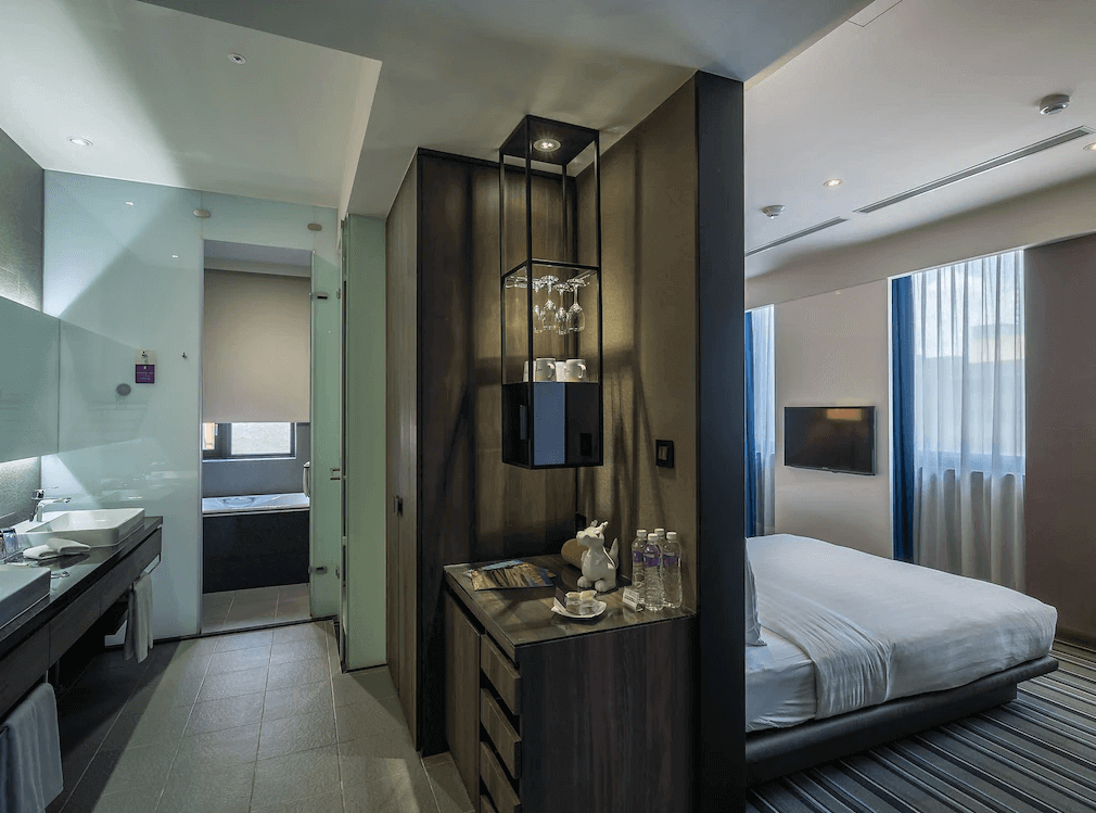 【西門町住宿】永安棧的開放式浴室和豪華套房，房內設有舒適雙人床，大片窗戶提供良好採光。房內設計融入深色木質元素，玻璃浴室隔間讓空間感更加開闊，盥洗用品與迎賓點心整齊擺放，提升住宿體驗。