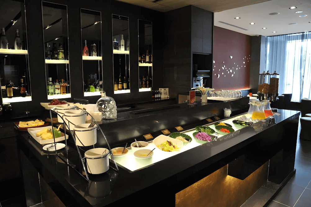 【西門町住宿】永安棧的餐飲區，提供豐富的自助早餐，包含沙拉、麵包、醬料、水果與果汁等。黑色大理石餐檯搭配溫暖燈光，背後的玻璃櫃展示各式酒品，營造高級且舒適的用餐氛圍。
