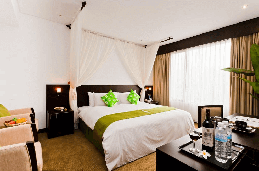 【金山溫泉】沐舍溫泉渡假酒店的精緻客房，配有一張大床，綠色抱枕與床尾巾增添自然氛圍。房內擺放了紅酒與礦泉水，並搭配柔和的窗簾與間接照明，營造出寧靜放鬆的住宿體驗。窗邊還設有休閒座椅與水果盤，適合享受悠閒時光。