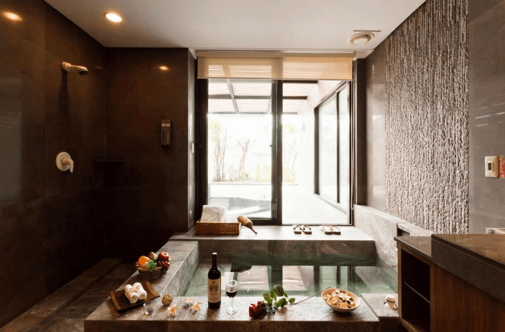 【金山溫泉】沐舍溫泉渡假酒店的高級溫泉浴室，配備寬敞的石材泡湯池，並擺放紅酒、水果與浴巾，增添奢華感。浴室牆面採用深色石材設計，搭配大片玻璃窗，提供戶外庭院的景觀，使泡湯時更加放鬆與私密。