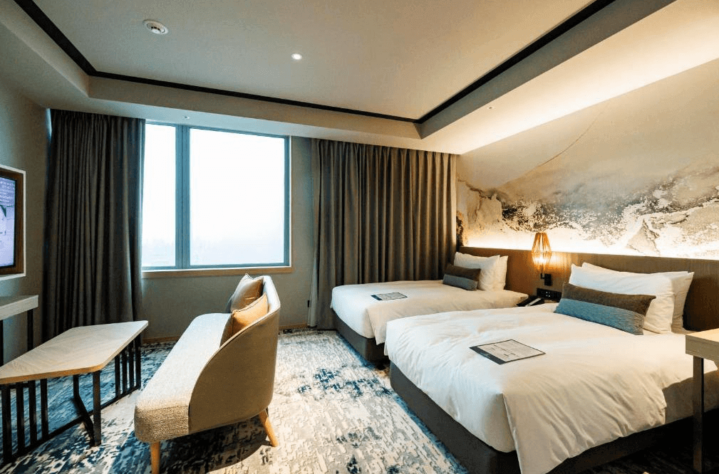 【西門町住宿】索拉利亞西鐵飯店台北西門的高級雙床房，擁有兩張單人床，搭配柔和的燈光與大面窗戶，自然光線充足。房間設計現代典雅，床頭牆面裝飾有藝術感的壁畫，並配有溫暖的床頭燈與舒適的座椅，營造出高級感與舒適的氛圍。