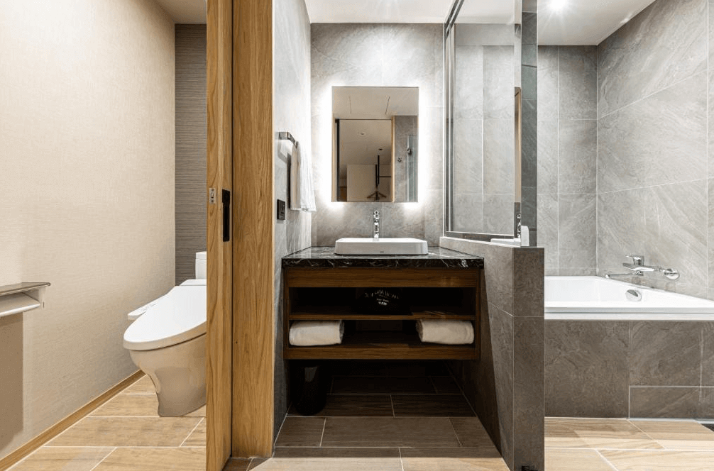 【西門町住宿】索拉利亞西鐵飯店台北西門的浴室，採用高級灰色大理石設計，營造簡約時尚感。浴室內有乾濕分離的設計，配有獨立浴缸、現代感鏡面與嵌入式燈光，洗手台設計精緻，底部櫃體提供收納空間，廁所區域則以木門區隔，確保隱私性。