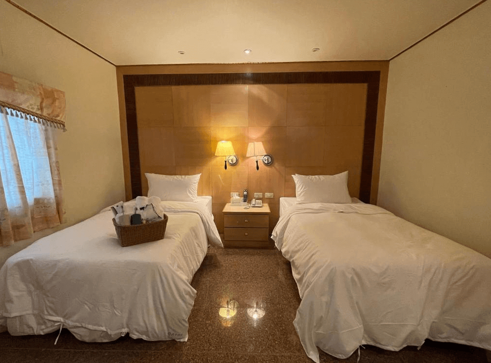 【四重溪溫泉】舍下立宇溫泉的雙床房，擁有兩張單人床，適合親友或旅伴入住。床頭牆採用淺色木紋設計，搭配壁燈與時鐘，增添典雅氣息。床頭櫃上擺放迎賓禮籃，窗戶搭配碎花窗簾，提升溫馨感，整體風格典雅大方。